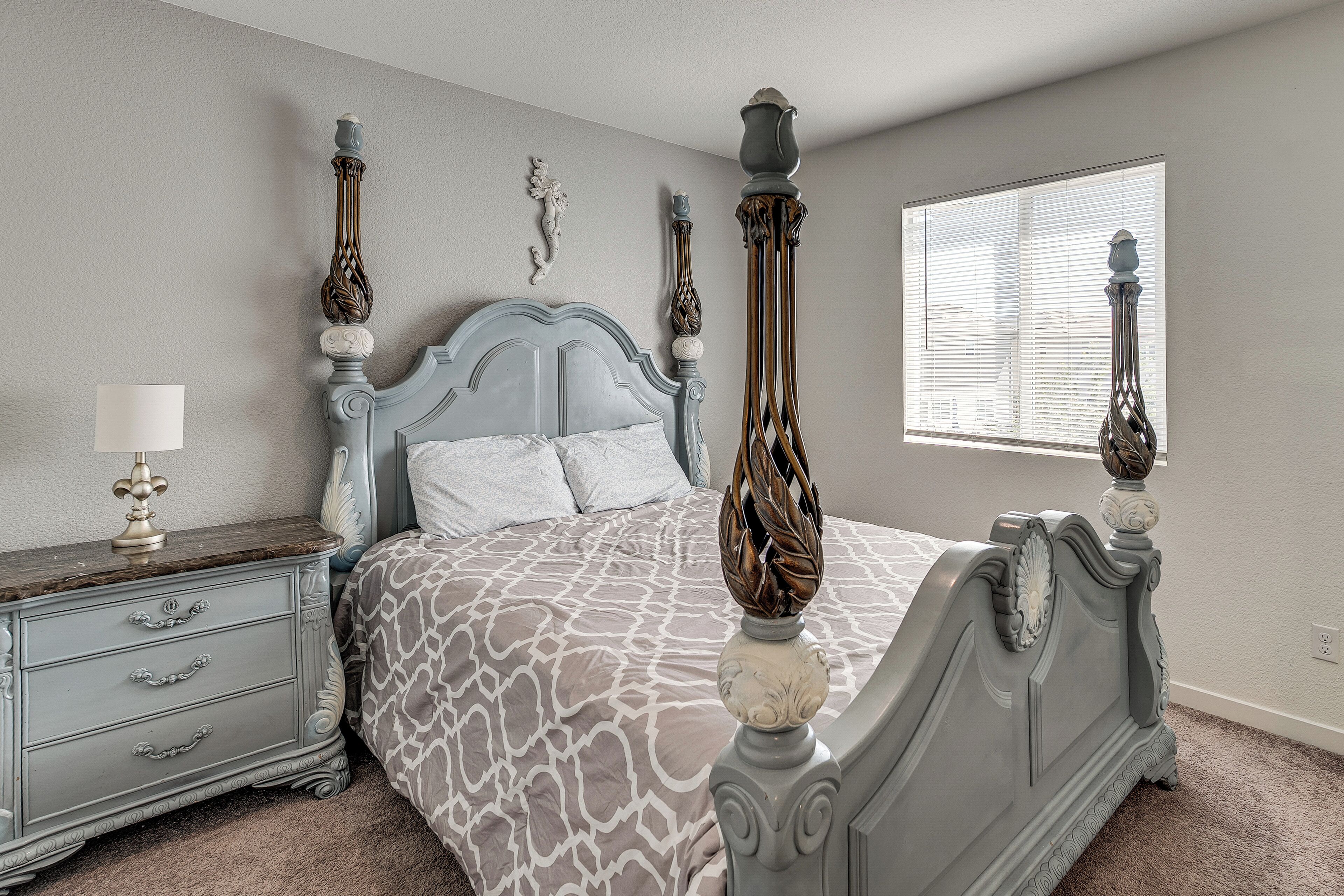 A bedroom with a four-poster bed, a nightstand, a lamp, and a window with blinds.
