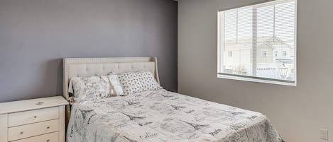Un dormitorio con cama, mesita de noche, ventana con persianas y vista a edificios.