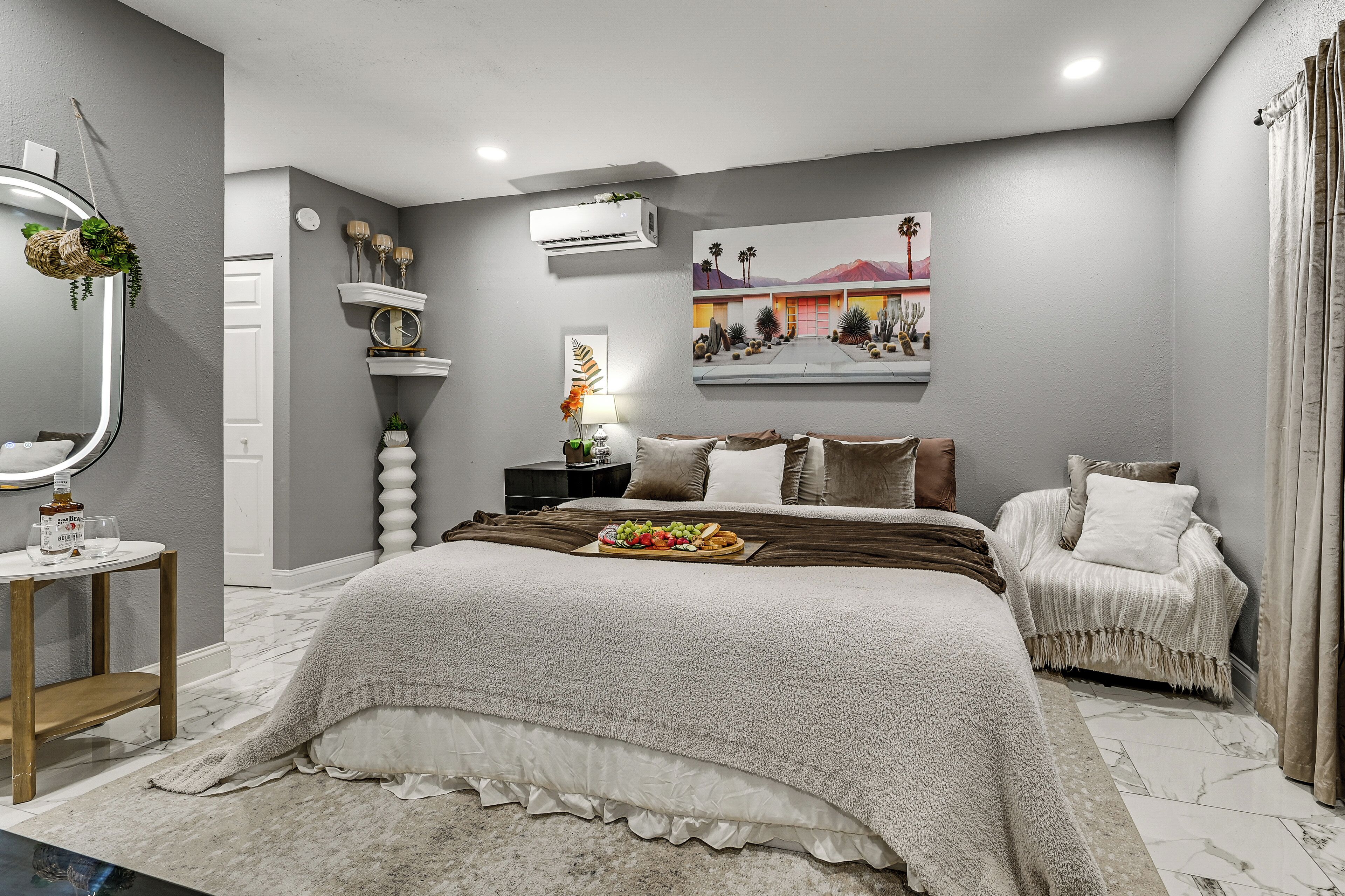 A bedroom with a large bed, a bedside table, a chair, a mirror, and a wall-mounted air conditioner.