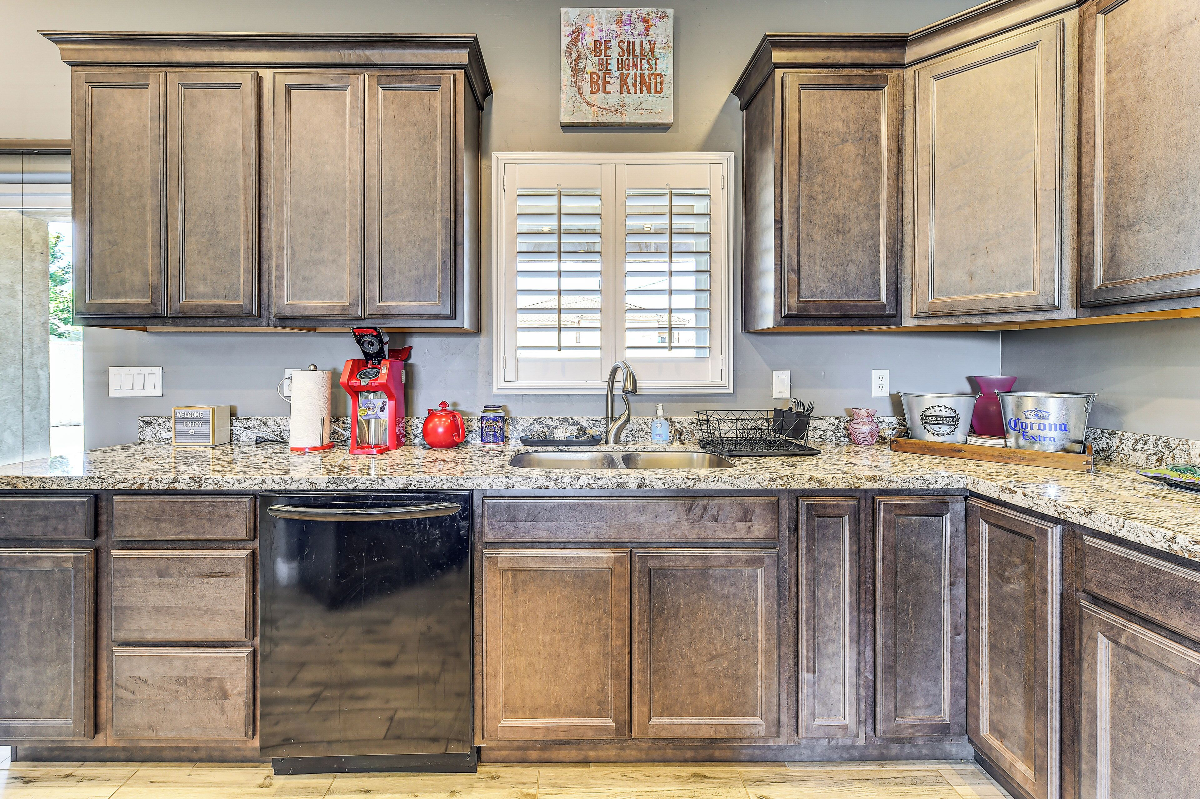 A kitchen with wooden cabinets, a built-in dishwasher, a window with blinds, and a countertop with various items.