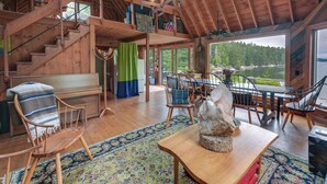 Ein geräumiger Loft-Raum mit hoher Decke, Holzbalken, einer Treppe, einem Essbereich mit Stühlen und Tisch sowie einem großen Fenster mit Blick auf ein bewaldetes Gebiet.