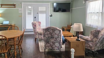 A living room with a wooden dining table, floral armchairs, a TV mounted on the wall, and a window with blinds.