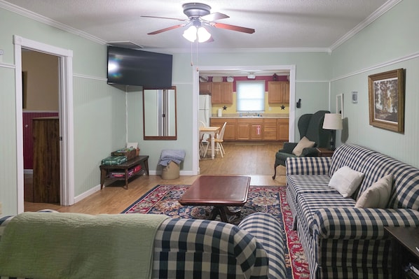 A living room with a sofa, a coffee table, and a TV. There is a kitchen in the background with wooden cabinets and a window.