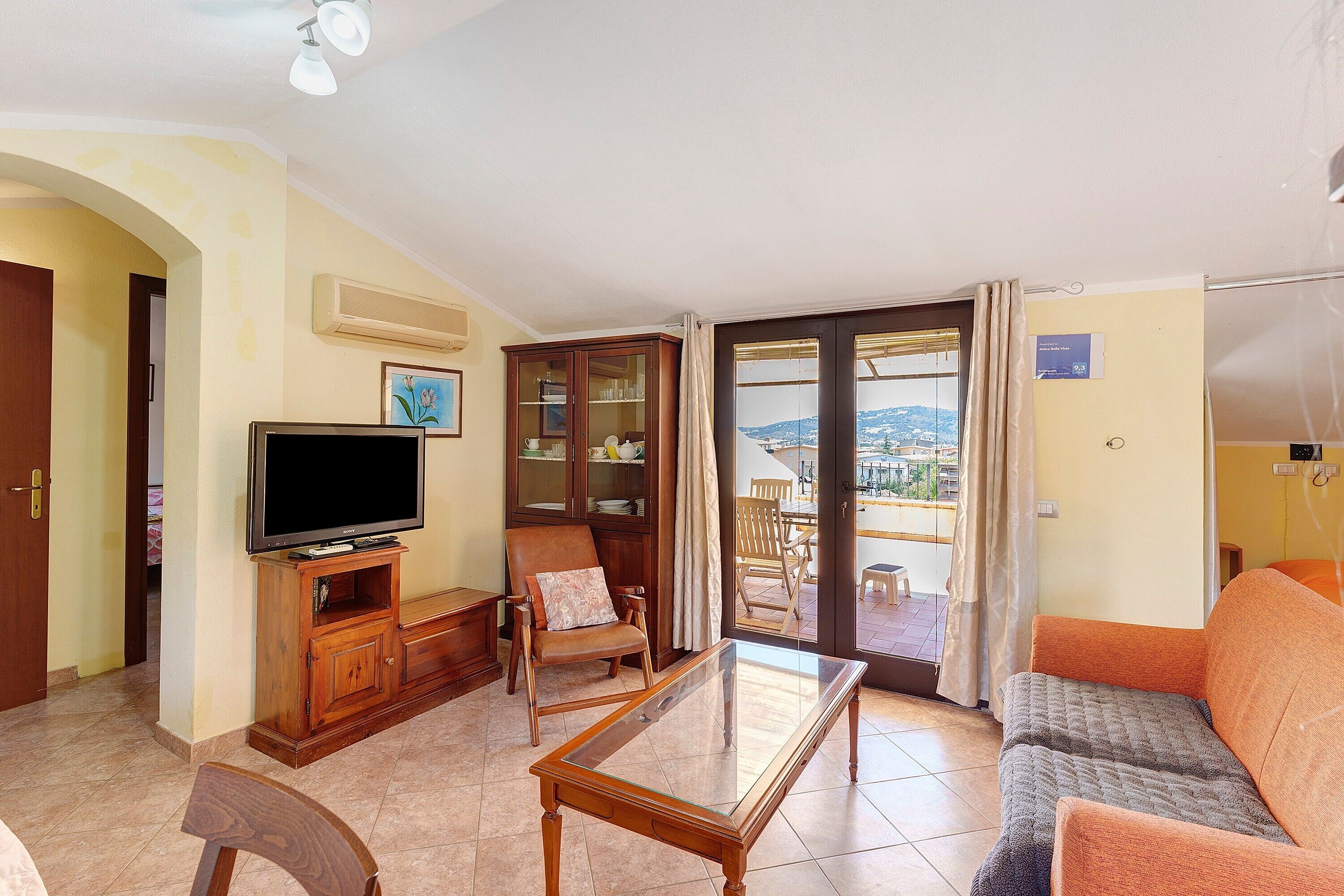 A living room with a television, a glass-top coffee table, a sofa, and a chair. There is a view of a balcony with a table and chairs outside.