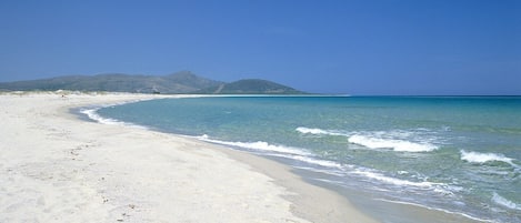 A beach with clear turquoise water, white sand, and distant mountains.