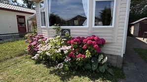 A small, single-story mobile home with a white exterior and a black roof. It features a large window with white curtains and a small porch area. - Kamperland Beach Resort - 1042 (Kamperland)
