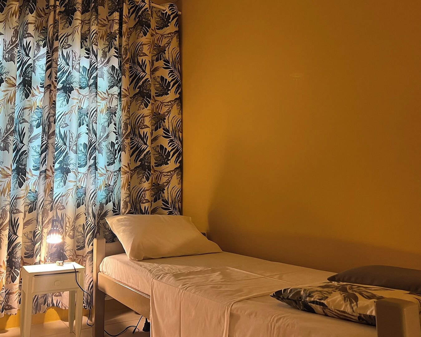 Ein Einzelbett mit weißer Matratze und Kissen, ein Nachttisch mit Lampe und ein blumiger Vorhang.