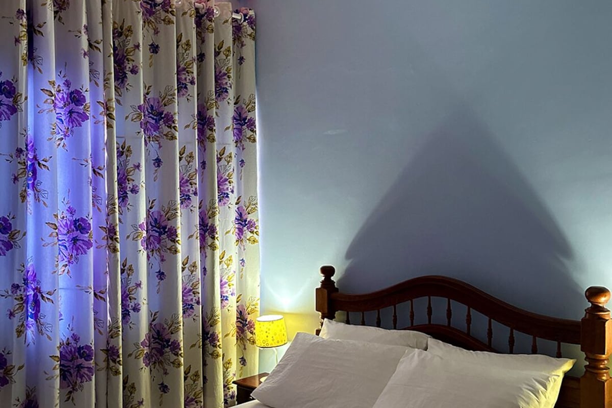 A single bed with white linens and a floral-patterned pillow, a wooden headboard, and a window with floral curtains.