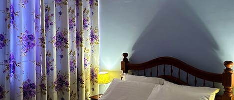 A single bed with white linens and a floral-patterned pillow, a wooden headboard, and a window with floral curtains.