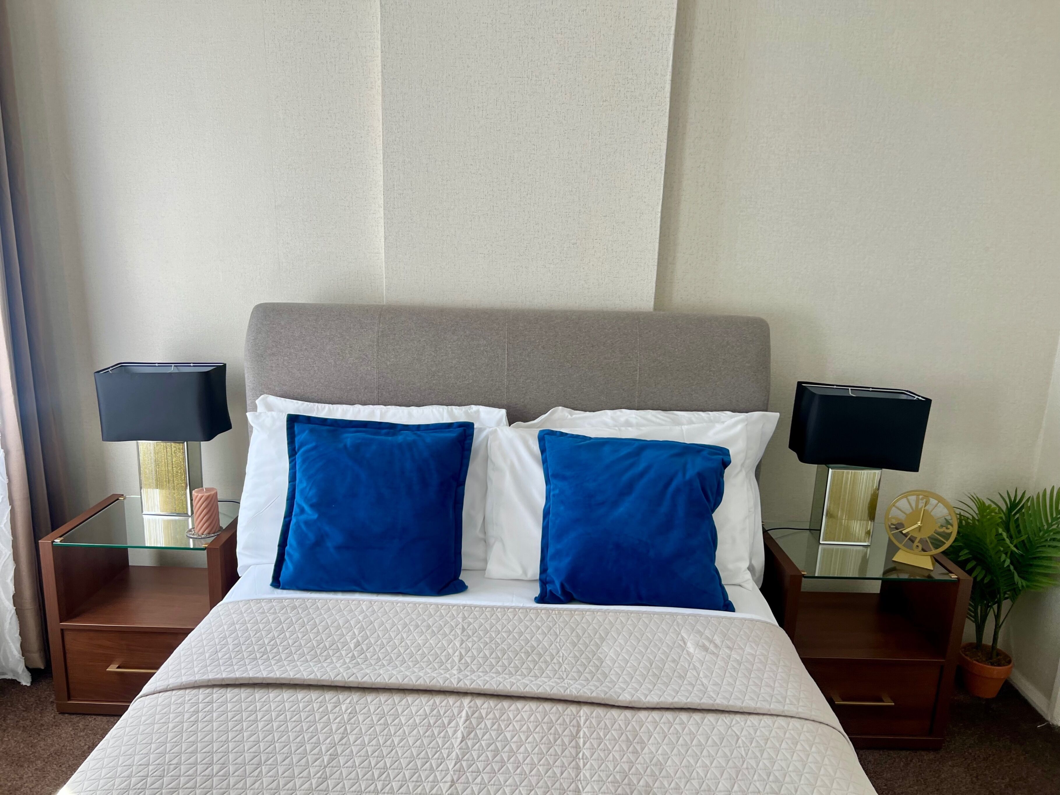 Una cama cuidadosamente arreglada con cubrecama acolchado, dos mesitas de noche con lámparas, una planta en maceta y un panel de pared con textura.