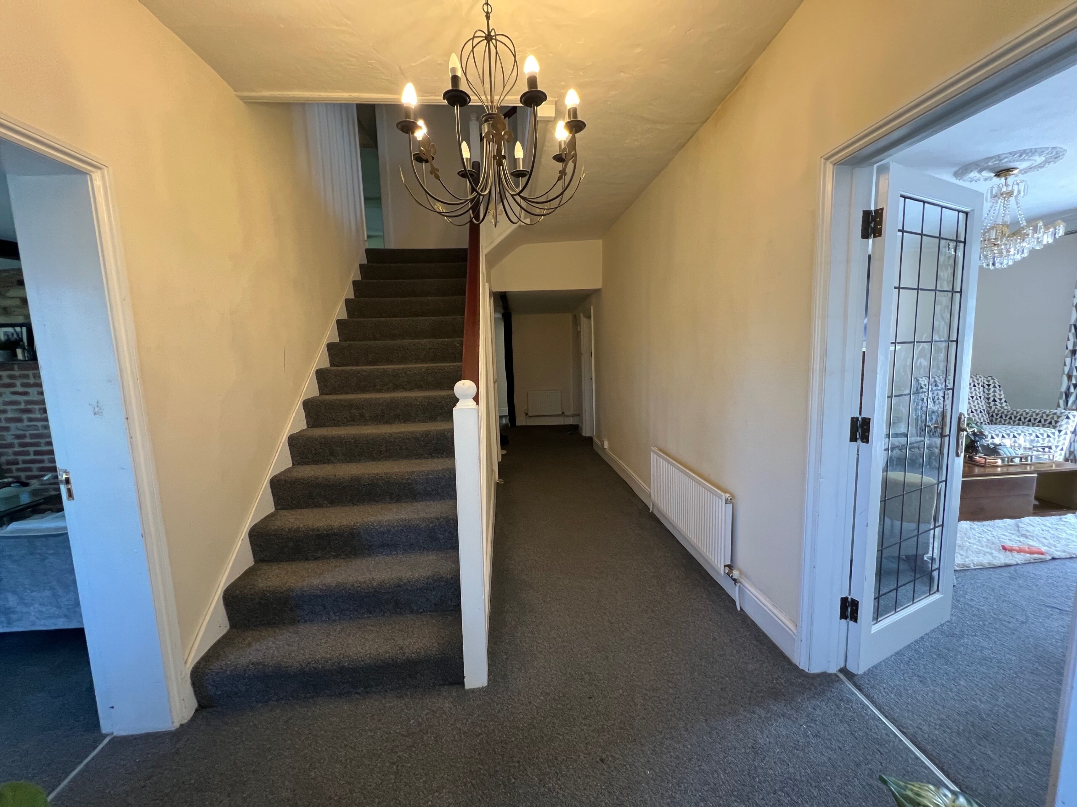 A carpeted staircase with a white handrail, a chandelier, and a door leading to a room with patterned furniture.