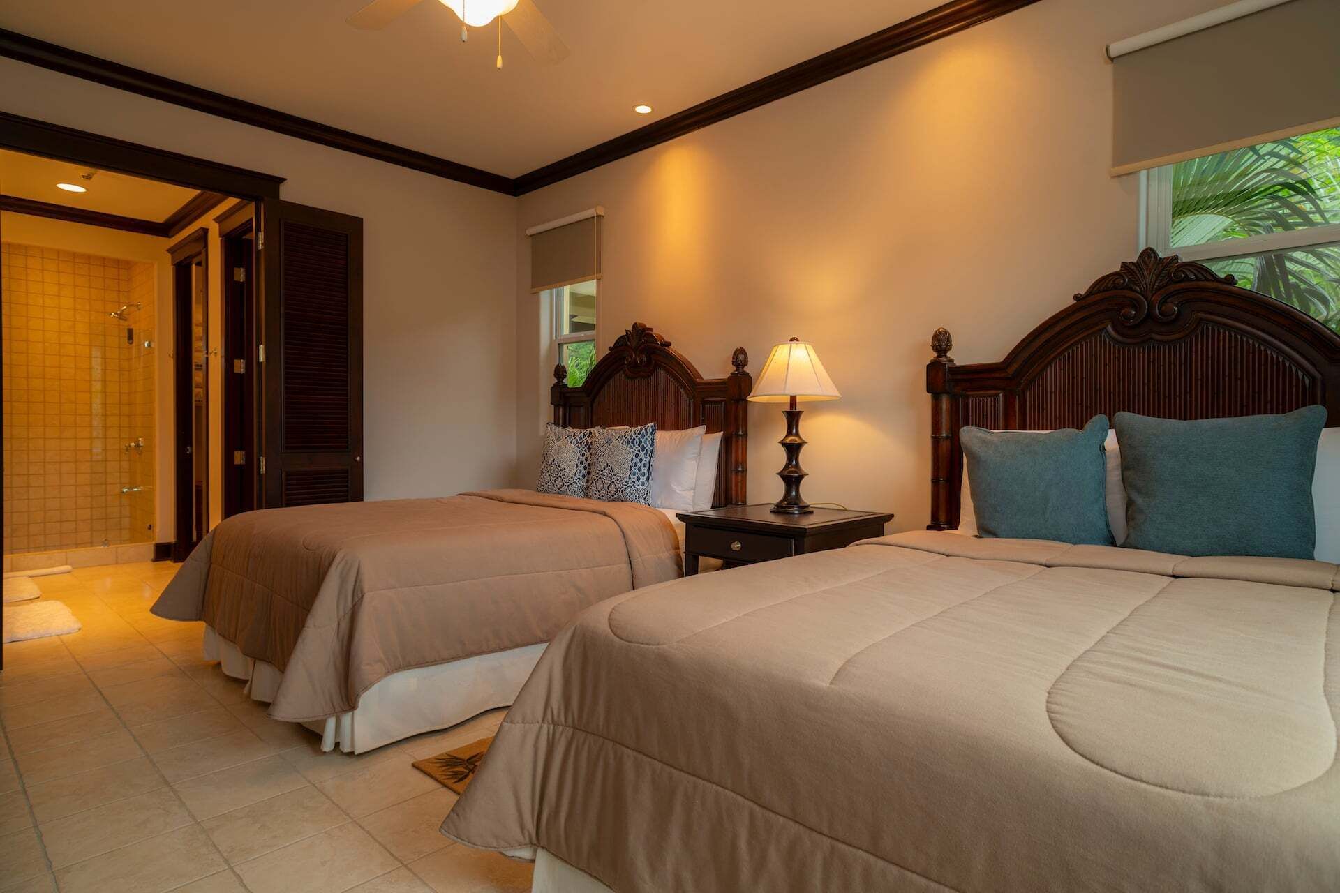 Habitación de hotel con dos camas, una mesita de noche con lámpara, baño con ducha y ventana con vista a zonas verdes.
