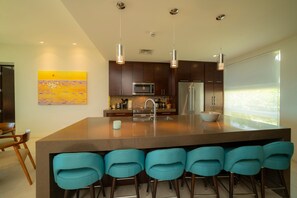 A modern kitchen with a central island, bar stools, stainless steel appliances, pendant lighting, and a large window. - Montecielo 4E 3 bdrs ocean view in Los Suenos (Herradura)