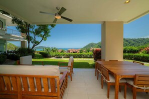 A patio with a wooden table and chairs, a ceiling fan, and a view of a lush garden and distant hills. - Montecielo 4E 3 bdrs ocean view in Los Suenos (Herradura)