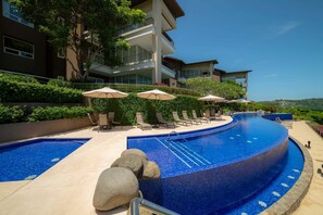 A modern hotel with a curved infinity pool, lounge chairs, and umbrellas. - Montecielo 4E 3 bdrs ocean view in Los Suenos (Herradura)