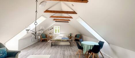 A loft-style room with a sloped ceiling, wooden beams, a bed, a dining table, and a sofa.