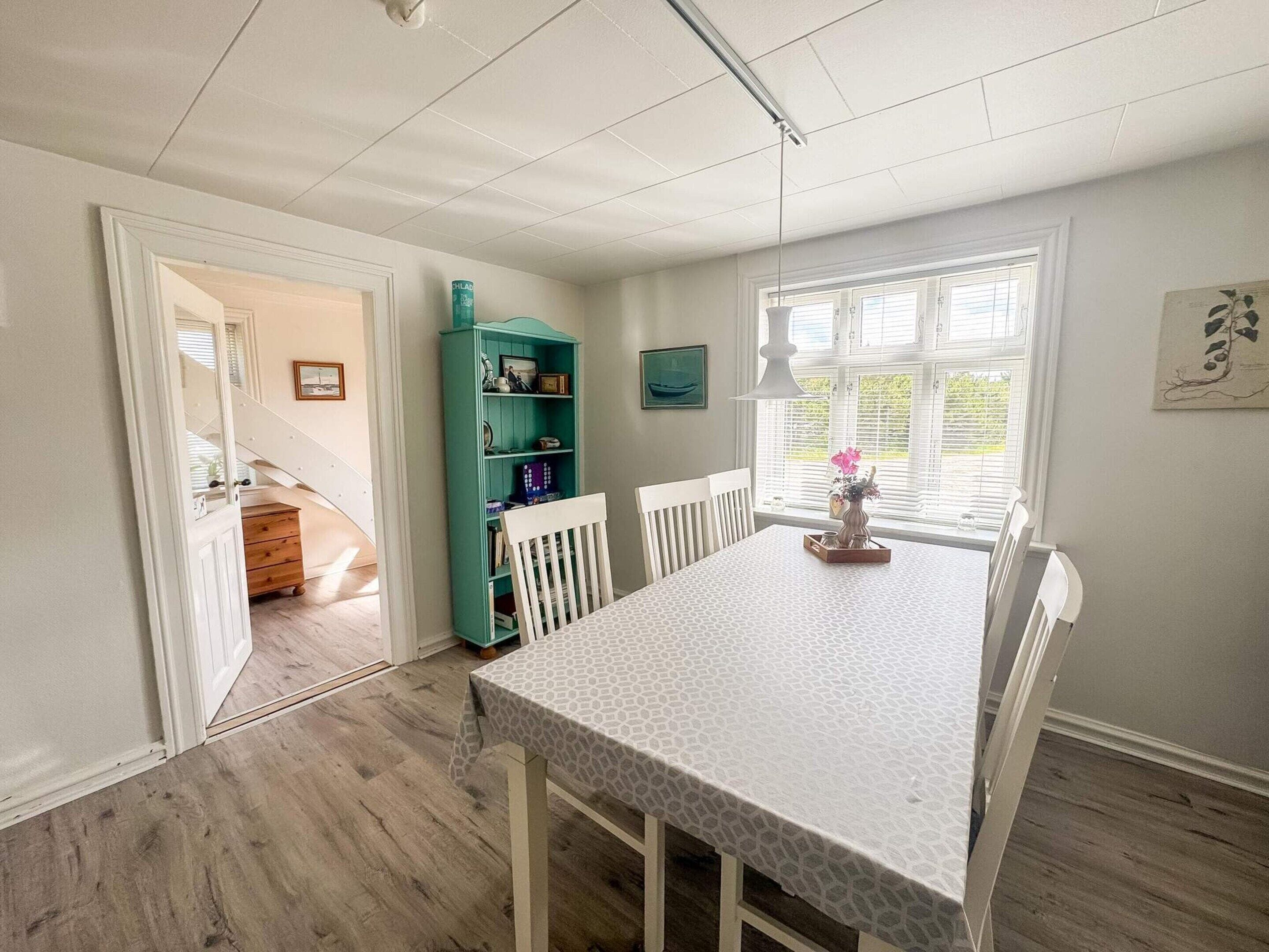 Un coin repas comprenant une table, des chaises, une bibliothèque de couleur turquoise et une fenêtre avec des stores.