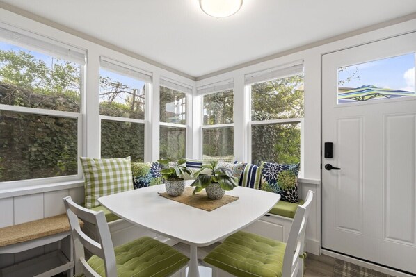 A sunlit room with a white table, green cushioned seating, and large windows.