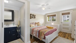 A bedroom with a bed, a ceiling fan, a window with a view, a door, and a bathroom with a sink and mirror.
