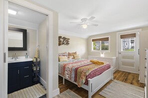 A bedroom with a bed, a ceiling fan, a window with a view, a door, and a bathroom with a sink and mirror.
