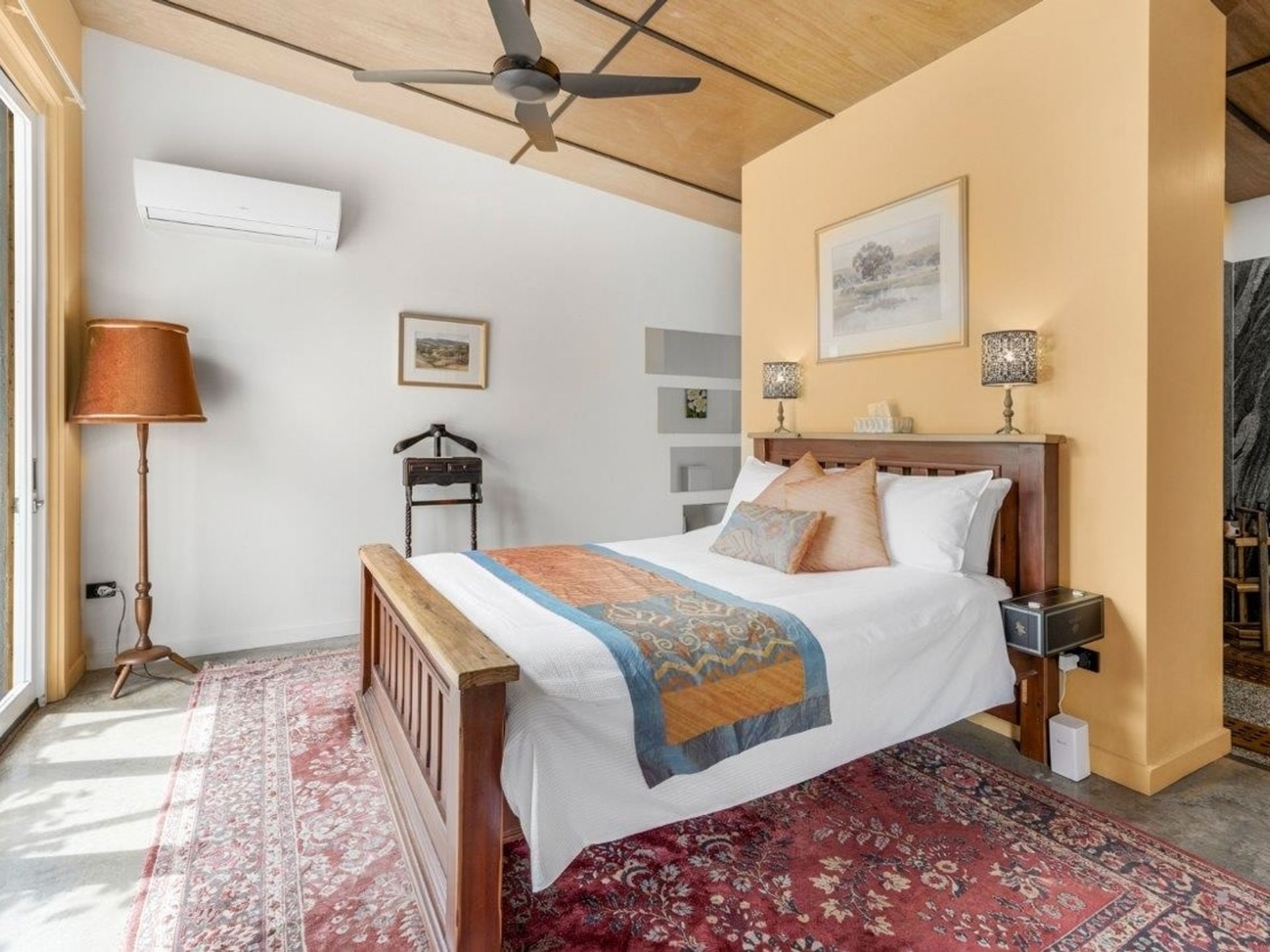A bedroom with a wooden bed, a red patterned rug, a ceiling fan, and a wall-mounted air conditioner.