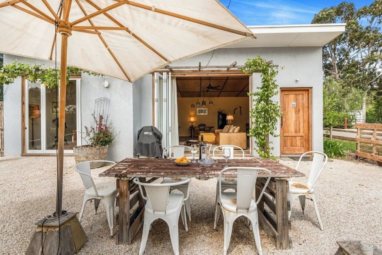 An outdoor dining area with a rustic wooden table and white chairs, an umbrella, and a view of the house interior.