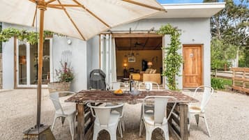Área de refeições ao ar livre com mesa de madeira rústica, cadeiras brancas, um guarda-sol e vista para o interior da casa.