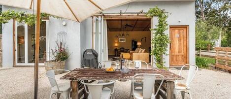 An outdoor dining area with a rustic wooden table and white chairs, an umbrella, and a view of the house interior.