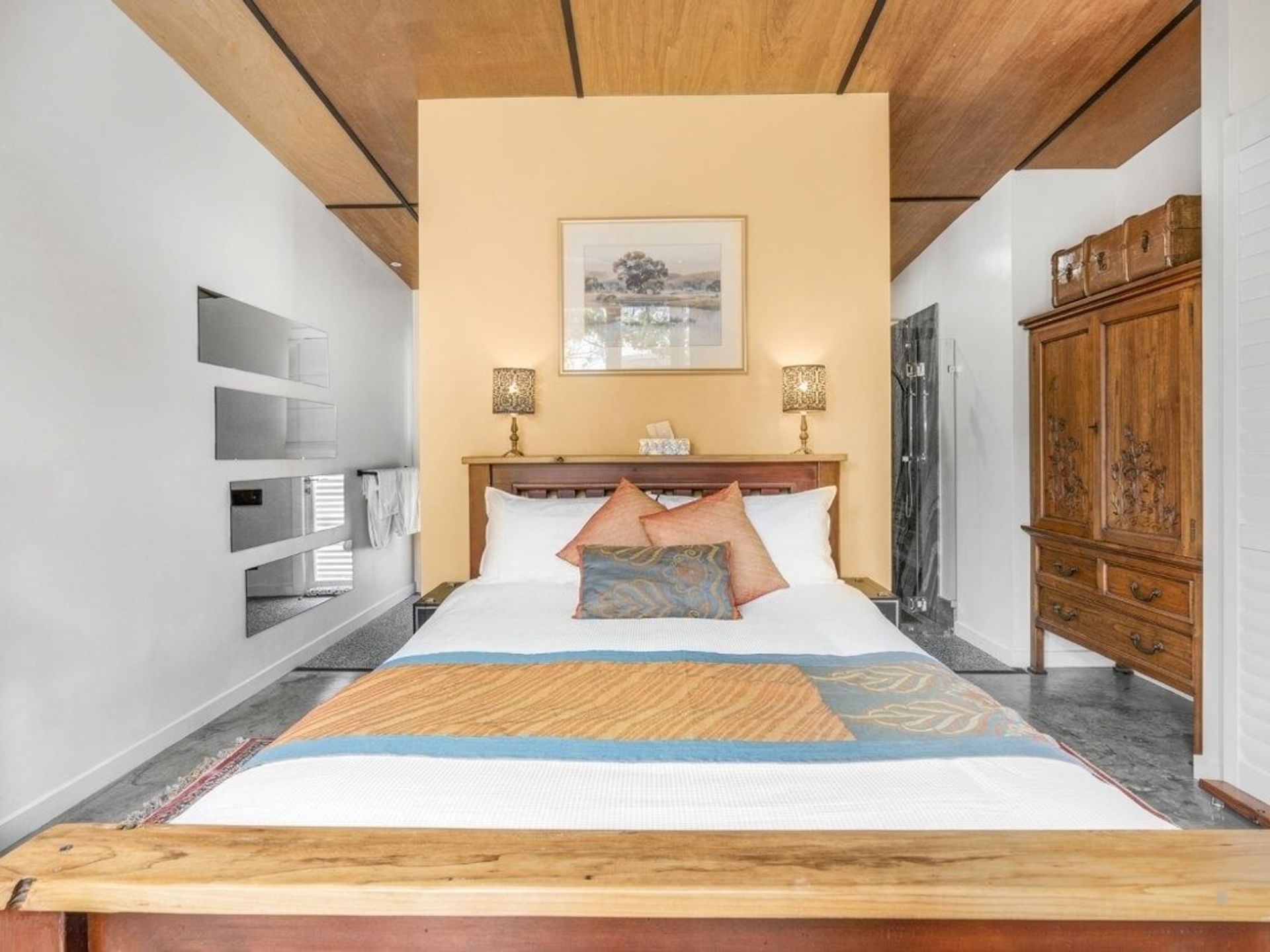 A hotel room with a large bed, wooden headboard, bedside lamps, a painting on the wall, and a wardrobe.