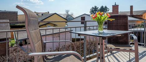 A balcony with a table and chairs, a vase with flowers, and a view of neighboring buildings.