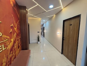 A hallway with a bench, a wall with a relief sculpture, a potted plant, and a door with a number. - Hotel Dev Shree Palace (Ujjain)