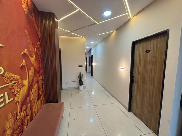 A hallway with a bench, a wall with a relief sculpture, a potted plant, and a door with a number. - Hotel Dev Shree Palace (Ujjain)