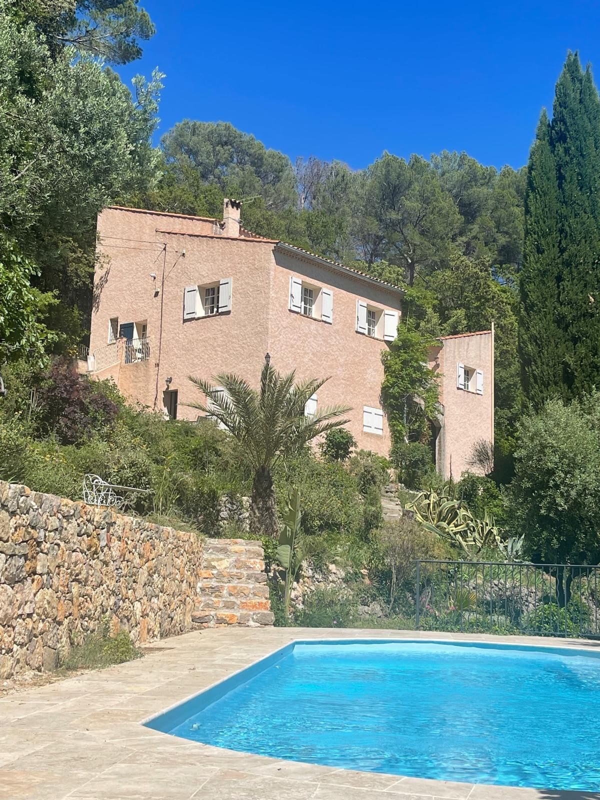 Un edificio de dos pisos con piscina, rodeado de follaje y un muro de piedra.