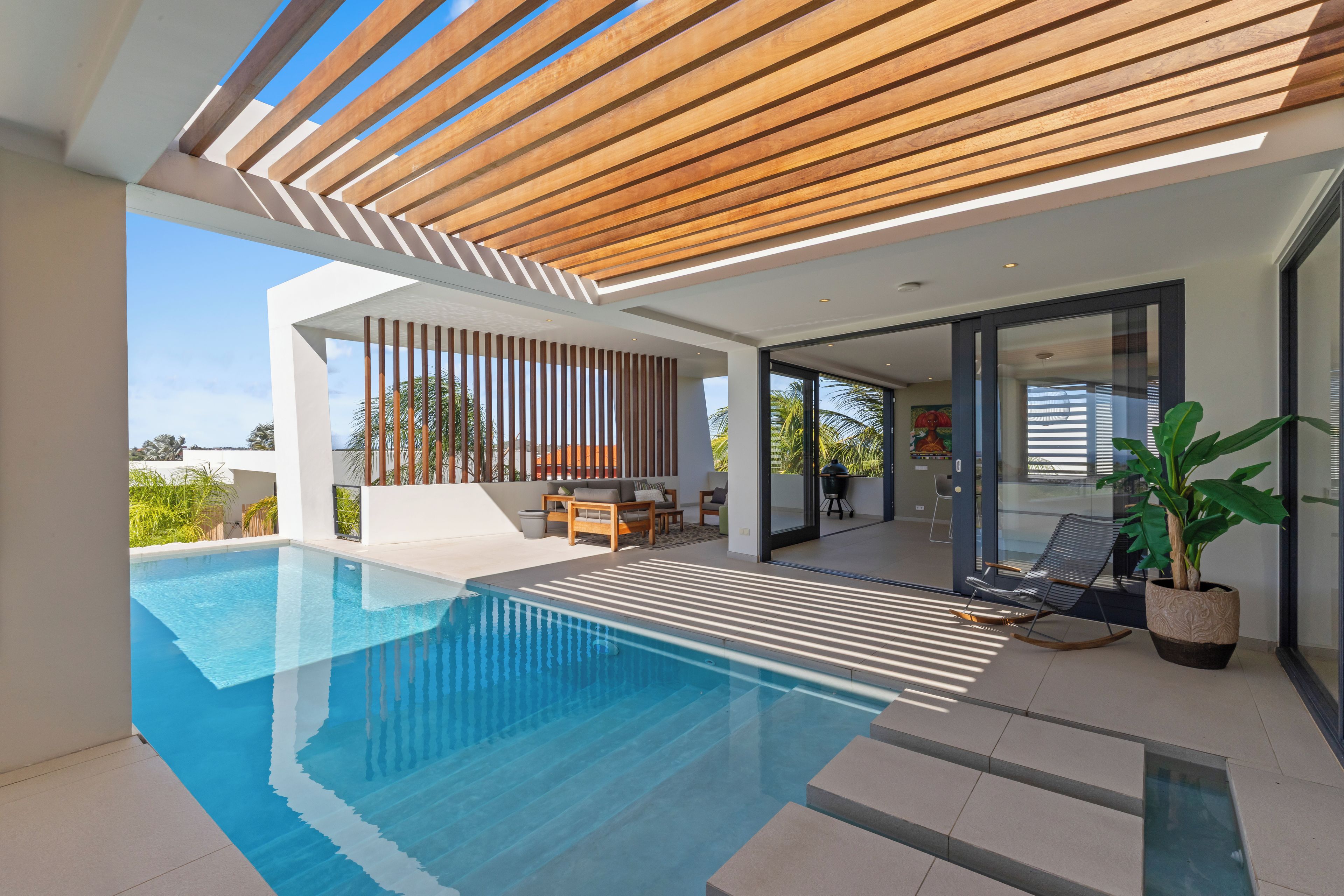 Un área de piscina moderna al aire libre con una pérgola de madera, un espacio de descanso con mesa y sillas, y una planta grande en maceta.