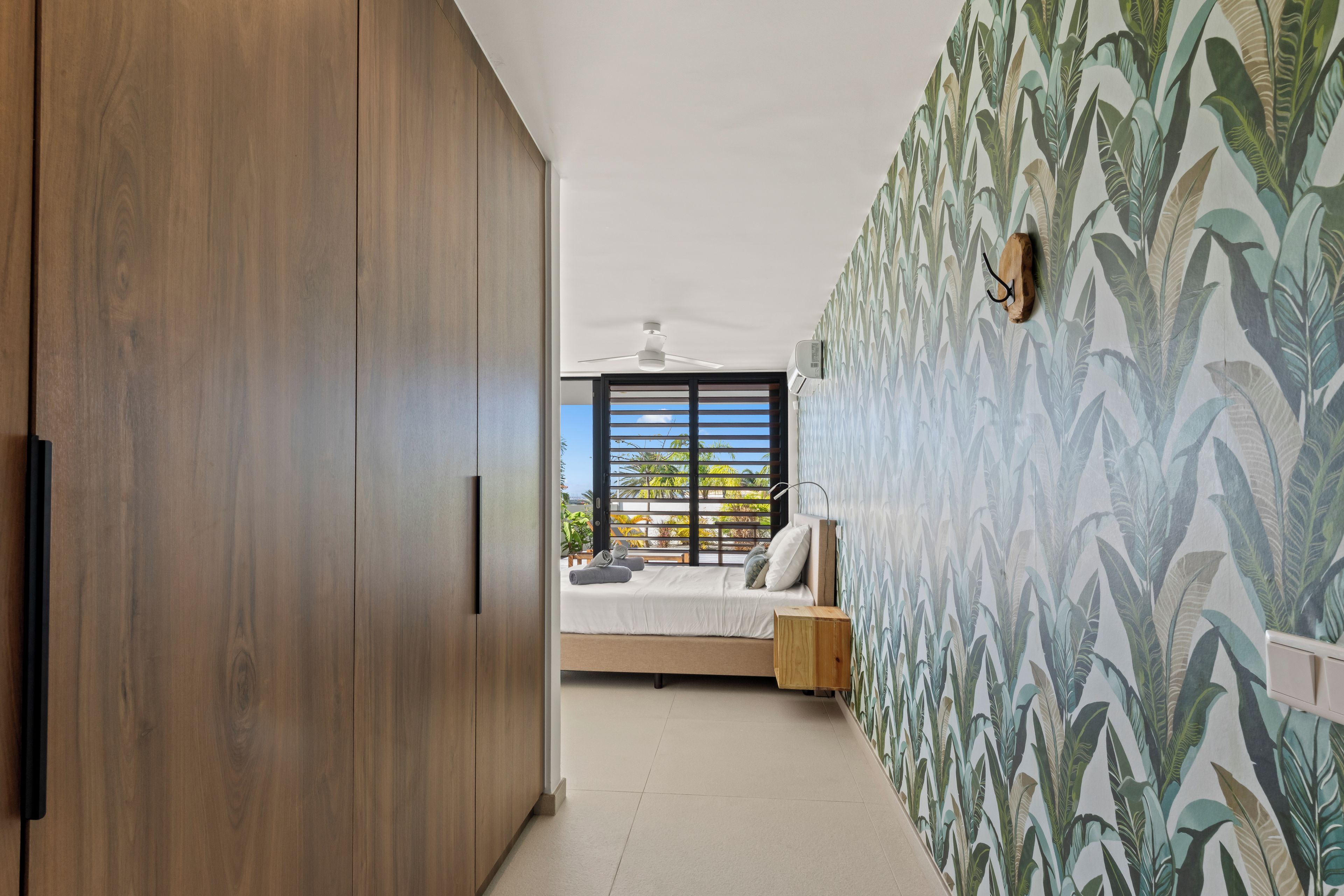 A bedroom with a bed, a wooden wardrobe, a ceiling fan, and a window with a view of greenery.
