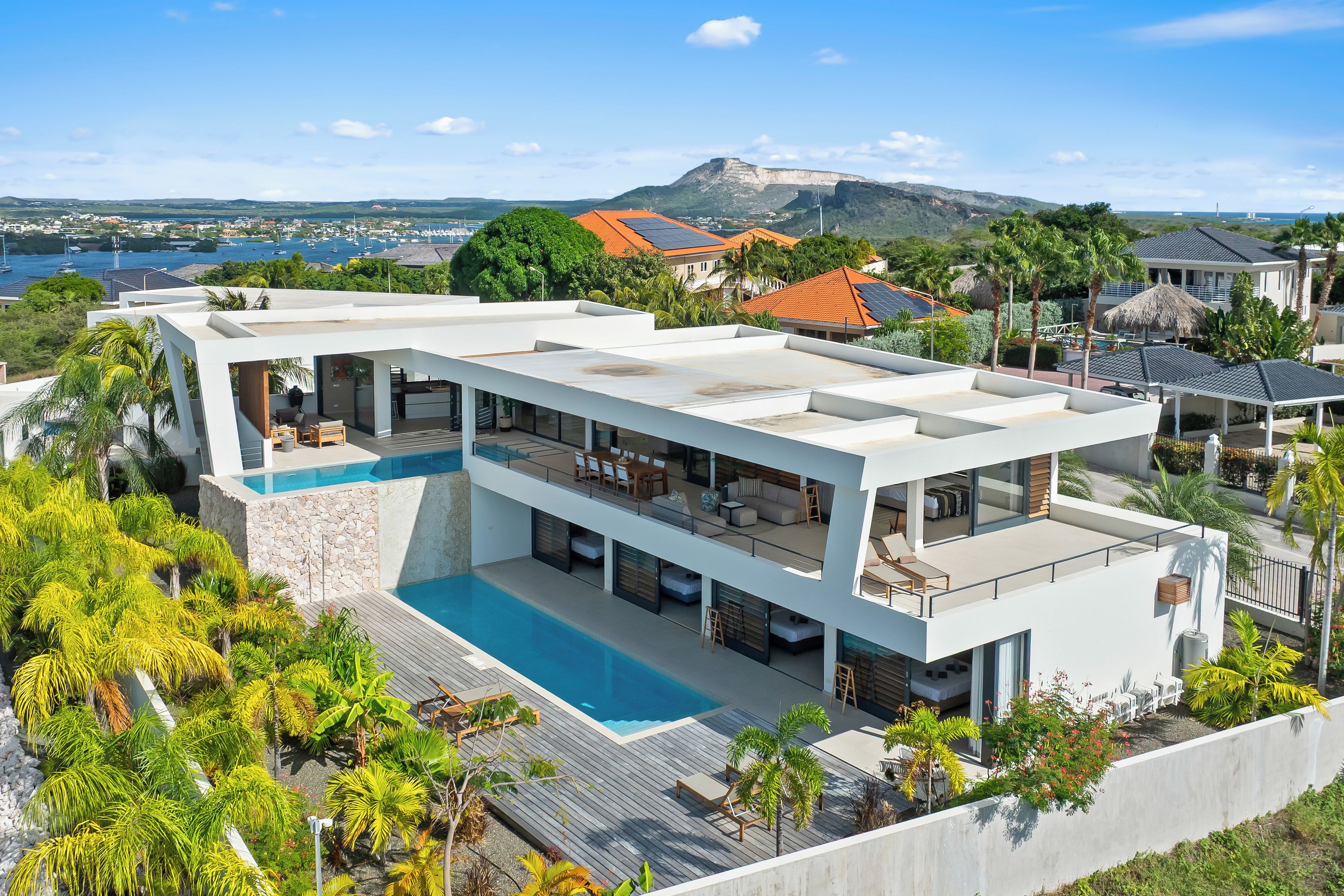 A modern multi-story house with a swimming pool, surrounded by lush greenery and a view of a mountain and the sea.
