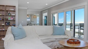 Ein modernes Wohnzimmer mit einem weißen Modulsessel, einem Holztisch und Blick auf das Meer durch große Schiebeglastüren.