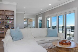 Een moderne woonkamer met een witte modulaire bank, een houten salontafel en uitzicht op de oceaan door grote glazen schuifdeuren.