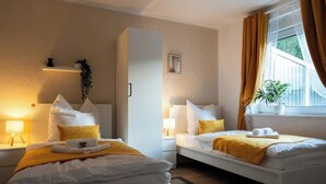 Quarto de hotel com duas camas, janela com cortinas, mesa de cabeceira com abajur e uma pequena prateleira com uma planta.