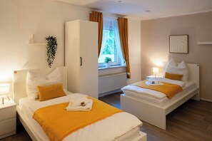 A hotel room with two beds, a wardrobe, a window with curtains, a bedside table with a lamp, and a wall-mounted shelf with a plant.