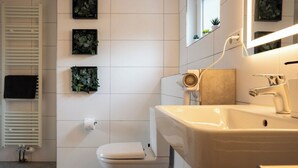 Casa de banho moderna com sanita branca, lavatório bege e espelho sobre o lavatório. Três plantas emolduradas na parede e um porta-toalhas à vista.