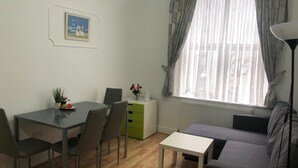 Ein Wohnzimmer mit einer Couch, einem Esstisch mit Stühlen, einem kleinen weißen Tisch, einem Heizkörper und einem Fenster mit Vorhängen.
