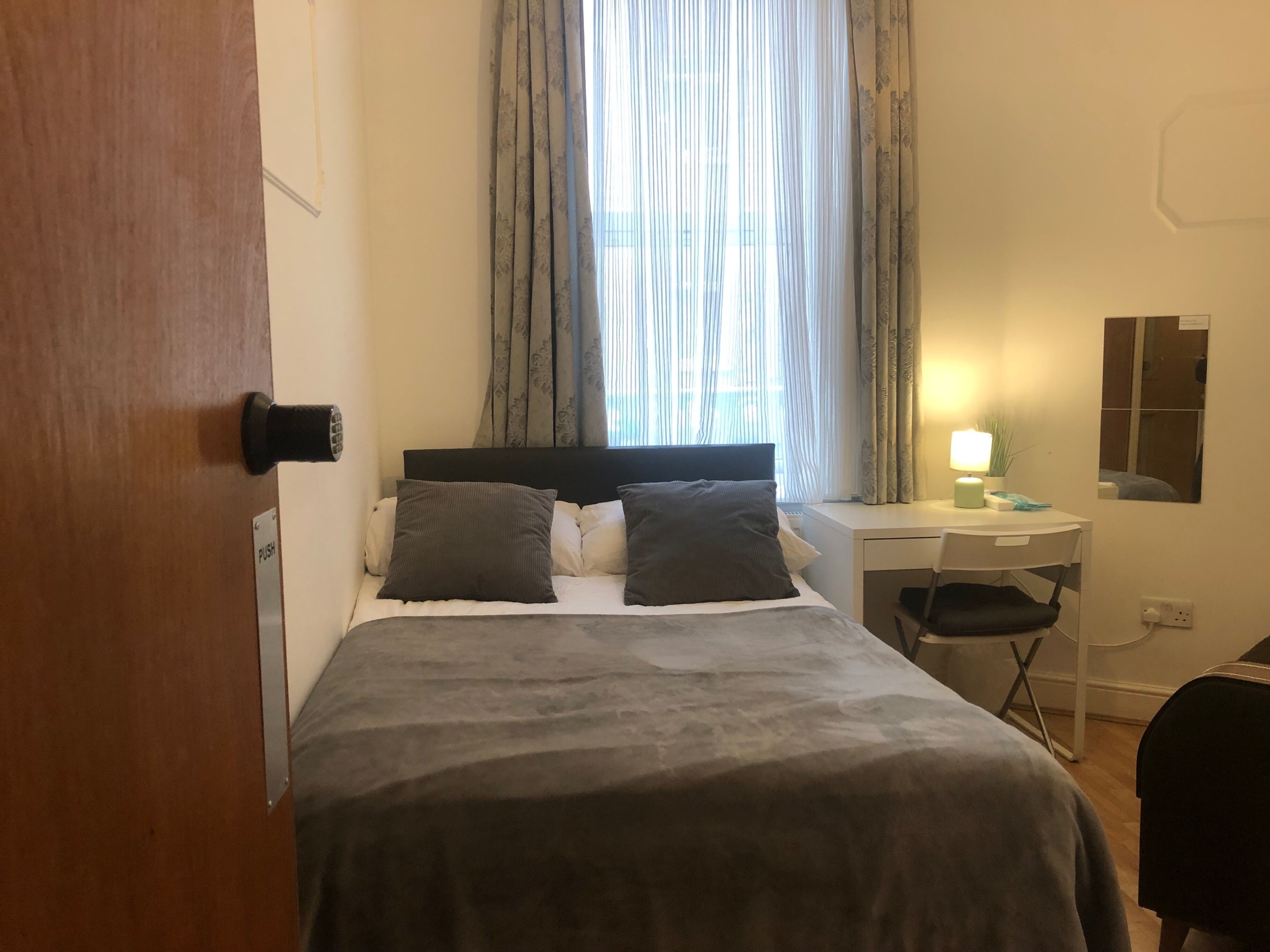 Ein Schlafzimmer mit einem Bett, einem Schreibtisch, einem Sessel, einer Lampe und einem Fenster mit Vorhängen.