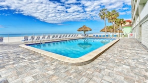 Ein Pool mit Liegestühlen und einem Strohdachschirm, eine Fliesenveranda und Blick aufs Meer.