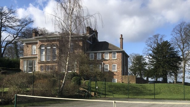 A tennis court with a net in the foreground, a brick building with multiple windows in the background, and a clear sky with some clouds. - Exclusive Apartment at Historic Hall With Hard Tennis Court (Tadcaster)