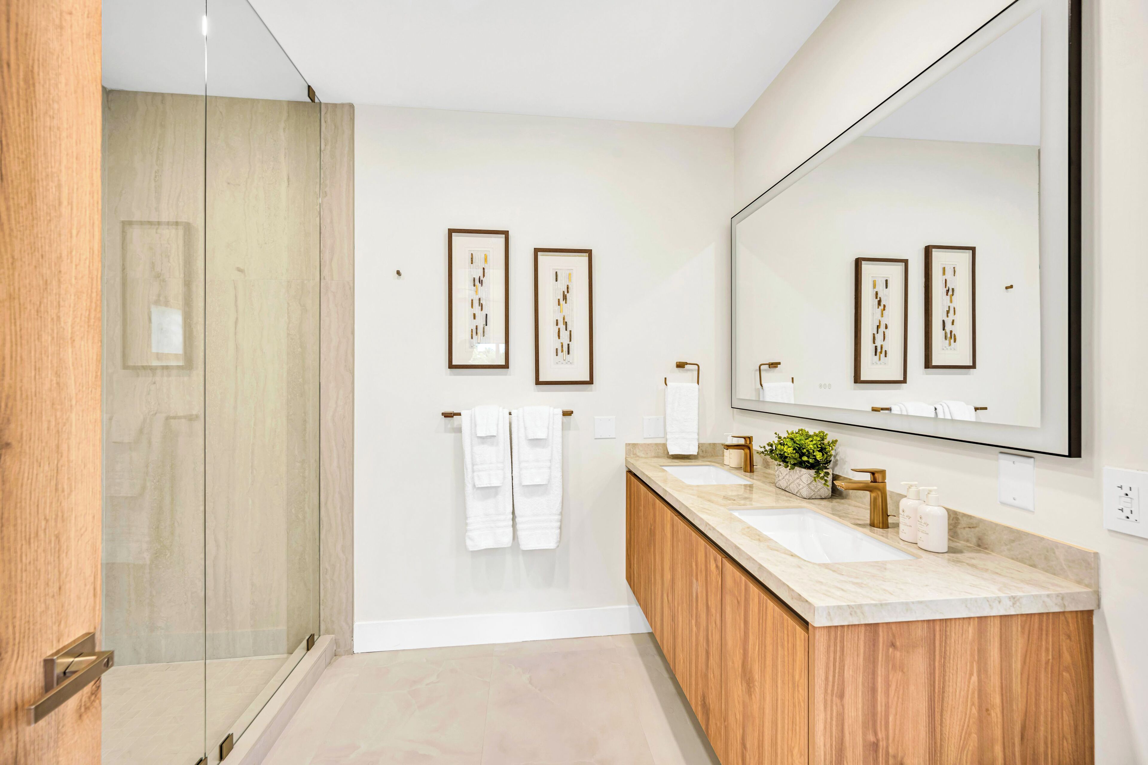 A modern bathroom with a glass shower, wooden vanity, two sinks, and framed artwork on the wall.