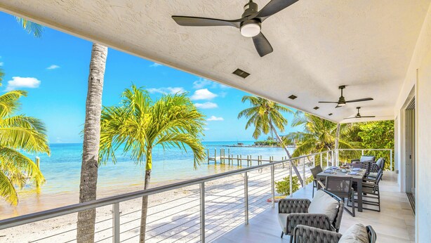 A balcony with a view of the beach and ocean, equipped with ceiling fans, outdoor furniture, and a railing.