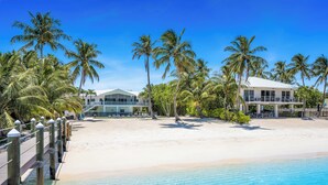 A beachfront property with a wooden deck, clear blue water, and palm trees.