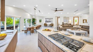 A modern kitchen with a central island, dining area with chairs, a living room with a sofa, and a ceiling fan.