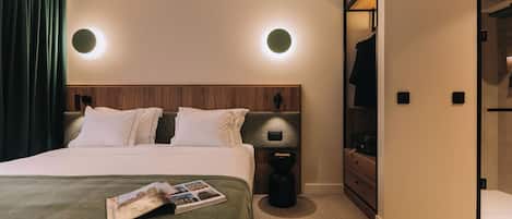 Una habitación de hotel moderna con una cama grande, cabecera de madera, dos lámparas de noche, una cortina verde, una revista sobre la cama y un vestidor con espejo.